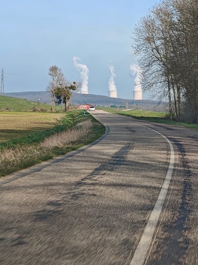 The image displays a rural landscape near Thionville, France, with the Cattenom Nuclear Power Plant visible in the background. A paved, two-lane asphalt road dominates the foreground, curving from the lower left towards the upper right. The road surface shows signs of wear and repair, and a solid white line marks its right edge. Long, dark shadows from unseen trees on the right stretch across the road. To the left of the road, a dry grass and dirt shoulder slopes upward to cultivated fields, presenting patches of bare earth and green growth.
In the midground, a white compact car is observed driving away from the viewer, navigating the road's curve. A blue and white road sign is visible on the left shoulder near the car. Further left, beyond the road, a small red-roofed building, possibly a barn or shed, stands on the edge of the fields. A bare deciduous tree, featuring prominent mistletoe clumps, is situated to the left of the red building. A metal lattice power line tower is discernible on the distant left hillside.
The background features a tree-covered hill range, behind which the four cooling towers of the Cattenom Nuclear Power Plant are prominently visible. Three of these towers are actively emitting large, white plumes of steam that rise into the clear, light blue sky, indicating operational status. The fourth tower, positioned to the far right, appears to be dormant or emitting negligible steam. No people are present in the scene.