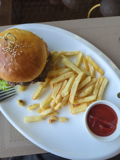 The image depicts a burger with sesame seed bun, accompanied by french fries and a small bowl of ketchup, presented on an oval white plate. A fork is positioned near the burger. The setting appears to be indoors, atop a woven placemat. No people are visible in the image. There is no specific indication within the image to confirm that it was taken in Chunkurchak, Kyrgyzstan.