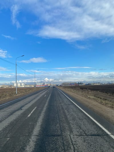 The image depicts a straight asphalt road stretching into the distance under a blue sky with scattered clouds. The road appears slightly wet, with visible reflections. To the right of the road is a dirt shoulder with vegetation. The left side features streetlights and a distant view of buildings, including what appears to be apartment complexes and industrial structures with smoke rising from chimneys. The location is identified as Usolye-Sibirskoye, Russia. There are a few vehicles in the distance along the road.