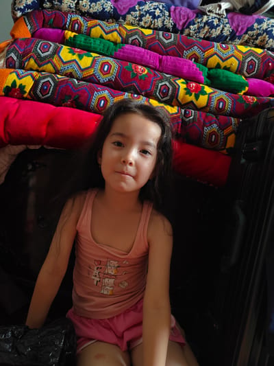 A young girl with long, dark hair sits in front of a stack of brightly colored, patterned textiles. The textiles are rolled and layered, creating a vibrant backdrop. The girl is wearing a pink tank top with a graphic of cats and the words "Let's Cat" and "hello!" visible on it, along with pink shorts. She is looking at the camera with a neutral expression. To the right of the girl, a black suitcase is visible. The scene appears to be indoors, with soft lighting. The colorful textiles suggest a cultural or domestic setting, possibly in Takhiatosh, Uzbekistan, as indicated by the location context. The girl's attire and relaxed posture suggest a casual moment, perhaps during a break or while relaxing at home. The overall impression is one of domestic comfort and cultural richness, highlighted by the distinctive textile patterns.