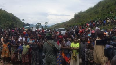 The image shows a large crowd of people gathered on a road in Sake, Democratic Republic of Congo. The crowd is positioned in the foreground, with a mountainous area, covered in lush vegetation, extending into the background on both sides of the road. The scene is set on a partly overcast day, with a bright sky above the crowd. The people are mostly dressed in casual clothing, and they appear to be standing calmly and peacefully. There are several vehicles parked on the road, including a truck, a van, and several cars. A man wearing a military uniform and green helmet stands in the crowd. A man with a camera is standing near the center of the scene. The camera is pointed towards the crowd. The image suggests that a public event or gathering is taking place, perhaps a meeting or rally.