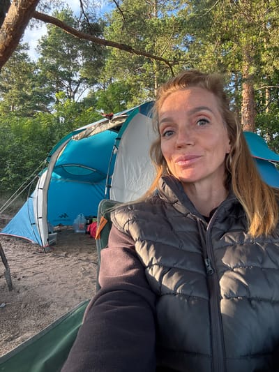 A woman is taking a selfie while sitting in a camping chair outdoors. She is wearing a dark puffer vest over a dark long-sleeved shirt and has blonde hair. Behind her, a blue tent is set up on sandy ground, surrounded by trees and vegetation, suggesting a natural environment. The lighting indicates it is daytime, possibly late afternoon or early morning, given the sun's angle. The image does not provide any specific details about the city of Shepelevo, Russia.