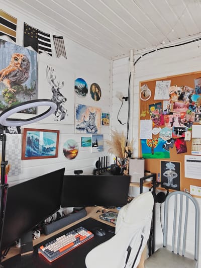 This is a medium shot, eye-level, indoor, interior photograph of a home office setup. The room is adorned with various paintings and decorations on a white paneled wall. In the center of the frame, a desk is equipped with two computer monitors, a keyboard, and a mouse. A white office chair is positioned in front of the desk. To the right of the desk, a bulletin board is covered with a collage of photos, drawings, and notes. The overall aesthetic suggests a creative and personalized workspace. The image does not provide any specific information about the city of Koledino, Russia, nor does it contain any people or depict any actions or interactions.