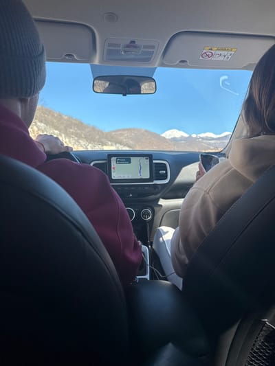 The image presents a forensic view from the rear passenger seat of a vehicle, looking forward at two occupants and the exterior landscape, presumably in the city of Šipačno, Montenegro.

**People:**
*   **Driver:** Positioned on the left, a male subject is seen from the back of the head and upper torso. He wears a grey knit beanie and a maroon/burgundy hooded sweatshirt. His right hand rests on the steering wheel, and a red string bracelet is visible on his left wrist.
*   **Front Passenger:** Seated on the right, a female subject is viewed from the back. She wears a light beige/tan hooded sweatshirt and white pants. Her right hand holds a dark-colored smartphone, which appears to be in active use.

**Vehicle Interior:**
The vehicle's interior is visible, featuring dark-colored front seatbacks. The dashboard is dark, equipped with a centrally mounted tablet-style infotainment screen displaying a navigation map. The screen shows a route with "4.0 mi" remaining and text that appears to read "Niksic Passage". Air vents with silver/chrome accents are present on either side of the screen. A gear shifter and charging cables are visible in the center console area. The rearview mirror reflects portions of the driver's head and the interior. Overhead, sun visors are visible, with the passenger-side visor displaying an "AIRBAG" warning label.

**Exterior Environment:**
Through the windshield, a clear, bright blue sky is visible. The landscape consists of rolling hills and mountains in the distance. Some hills in the foreground exhibit sparse brown vegetation and patches of snow, while prominent snow-capped mountain peaks are visible further in the background, indicative of a mountainous region. The scene is illuminated by daylight.