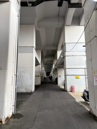 A narrow alleyway with large, white, rectangular ductwork lining both sides. The ducts are stacked and protrude into the space. The asphalt pavement is visible on the right. People are walking in the distance, under the ducts. A gray pipe runs along the bottom left. Stickers and signs are attached to the ductwork. A motorcycle is parked on the right, at the far end. The lighting is dim, with occasional spotlights on the ducts.