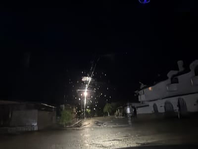 The image shows a nighttime scene in Umarevtsi, Bulgaria, likely captured during or after a rain shower. The foreground is dominated by a wet, paved road reflecting the light. A central street light illuminates the area, casting a bright glow and highlighting the falling rain. Structures line both sides of the road, including a white building on the right with arched windows and an irregular roofline, and a lower, darker building on the left. The overall environment is dark and wet. There are no visible people or interactions captured in this image.