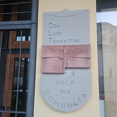 This is a medium shot of a memorial plaque mounted on a cream-colored building exterior in Calcinate, Italy. The plaque is made of grey stone with carved lettering and a pink stone artistic element. The top section of the plaque reads "DON LUIGI TENGATTINI". Below this is a horizontal pink stone carving that resembles an open book or a folded document with decorative clasps. The bottom section of the plaque is a grey stone semi-circle with more carved lettering that reads "SALA DELLA COMUNITÀ". To the left of the plaque, a dark grey metal gate with vertical bars is visible, obscuring part of a wooden door or window frame behind it. A large window is to the right of the plaque, reflecting the sky and buildings across the street. The overall impression is one of a public building or institution.