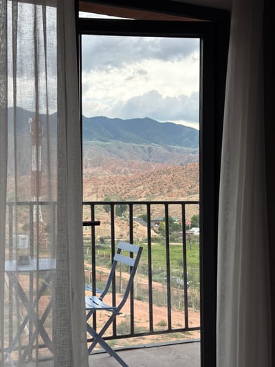 The image shows a view from a balcony overlooking a valley in Kaji-Say, Kyrgyzstan. The foreground features a light blue metal folding chair and a small table, partially obscured by sheer white curtains on the left and right sides of the frame. A metal railing separates the balcony from the view beyond. In the mid-ground, there are scattered buildings with colorful roofs, green grass, and dirt paths. A tall, red and white tower, possibly a communication tower, is visible on the left side, seen through the sheer curtain. The background reveals a dramatic landscape of layered, eroded mountains with distinct red and green hues under a cloudy sky. The overall scene depicts a rural or semi-rural setting with a striking natural backdrop.