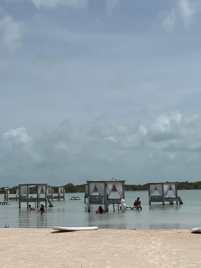 The image captures a daytime scene at a shallow bay or lagoon in San Pedro Town, Belize, under an overcast sky with patches of brighter clouds. In the foreground, a sandy beach occupies the lower portion of the frame, featuring a white surfboard lying flat on the sand and a blue container to its left. A pair of dark flip-flops rests on the sand further into the scene.

The midground is dominated by a body of calm, light blue-green water containing multiple elevated wooden structures resembling cabanas or gazebos, each draped with white curtains. These structures are built on stilts and are partially submerged in the shallow water.

From left to right within the water:
- The leftmost visible structure has two adults and one child in the water around it; the child, wearing orange shorts, stands in the water. This structure includes a small slide entering the water.
- To the right, another structure is partially visible.
- Centrally, a structure hosts three individuals. One person, dressed in white, stands in the water next to a table within the structure, appearing to interact with two others seated at the table.
- Further to the right, a person is partially submerged in the water next to another wooden structure.
- Additional similar structures are visible further back in the water.
- Several smaller, simple wooden tables or benches are dispersed in the water between the main structures.

The background consists of a dense line of green vegetation, likely mangroves, bordering the water and extending towards the horizon.