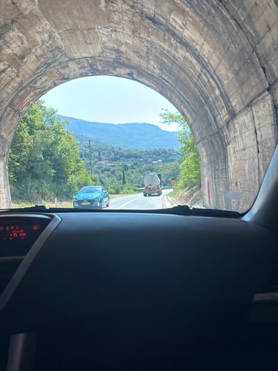 The image captures a view from inside a vehicle exiting a tunnel. The tunnel is constructed of rough, arched concrete with visible markings and texture. Outside the tunnel, a bright, sunny day reveals a lush green mountainous landscape. A paved road curves through the scenery, with trees and vegetation lining its sides.
On the road, a blue car is visible in the foreground, followed by a white tanker truck further in the distance. The dashboard of the vehicle taking the photo is visible at the bottom, displaying information including a speed of 115 km/h. The location is identified as Prijevorac, Montenegro.