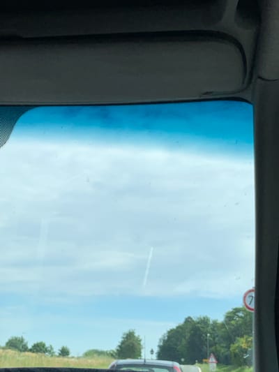 The image is taken from inside a vehicle, looking through the windshield. A dark gray sun visor is visible at the top of the frame. The windshield has a blue tint at the top and shows some dirt or smudges. Outside, the sky is bright and cloudy, with what appears to be a contrail from an airplane. In the distance, there are trees and vegetation. A circular speed limit sign with the number "70" inside a red circle and white background is visible on the right side of the frame. A triangular road sign is also visible in the distance. The bottom of the image shows the dashboard or part of the car interior, and the rear of another vehicle is partially visible. Based on the presence of the speed limit sign, the scene appears to be on a road in or near Oberwetz, Germany, a rural area known for its speed limits.