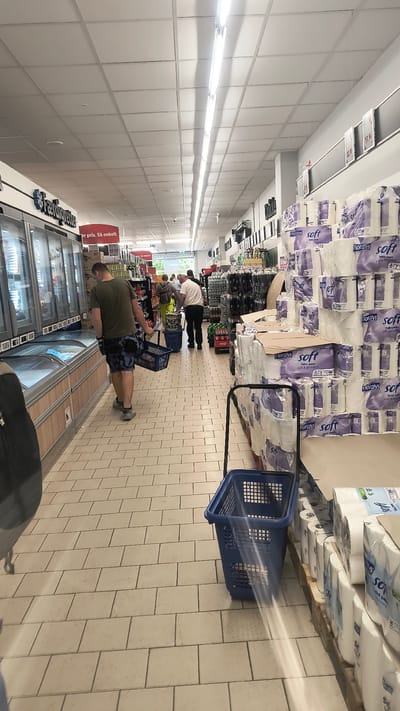 The image shows a vertical shot inside a supermarket aisle in Ølby Lyng, Denmark. The aisle is lined with shelves displaying products, primarily large packs of toilet paper in purple and white packaging. The flooring is tiled with light-colored square tiles. Fluorescent lights illuminate the scene from the ceiling.

Several people are visible shopping. In the foreground, a person wearing a green t-shirt and camouflage shorts is looking at a refrigerated display case. Further down the aisle, another person in a white shirt and dark pants is pushing a shopping cart. Other shoppers are present in the background, some appearing to be browsing or waiting. Two blue shopping baskets are prominent in the middle of the aisle.

Visible text includes "Floralys soft Toilet paper" on the product packaging and "er pris. Så enkelt" on a sign, which translates to "at a price. So simple". The overall impression is a typical grocery shopping experience. The lighting suggests it is daytime.