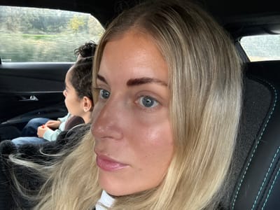 The scene depicts two individuals inside a vehicle. The user-provided context places this scene in Pourrières, France; however, this specific geographic location is not ascertainable from the visual content alone.
In the foreground, a female subject with blonde hair and light blue eyes is partially visible, oriented slightly towards the right of the frame. Her right eyebrow exhibits dark, well-defined hairs, contrasting with her lighter hair. Her lips are light pink. She appears to be seated in a front vehicle seat, with a black seat featuring teal stitching visible to her right. A white garment is partially visible on her person.
In the mid-ground, a younger individual with dark, curly hair pulled back is seated in the rear passenger seat, observed in profile. This individual is looking out the left rear window. They are wearing a light blue or mint-colored long-sleeved top and dark blue trousers. Red nail polish is visible on one of their fingernails as their hands are clasped.
Through the blurred rear window, an exterior environment is visible, characterized by green foliage and a reflective surface, likely water or a wet road, under what appears to be an overcast sky. The vehicle's interior paneling, including a door handle, is dark.