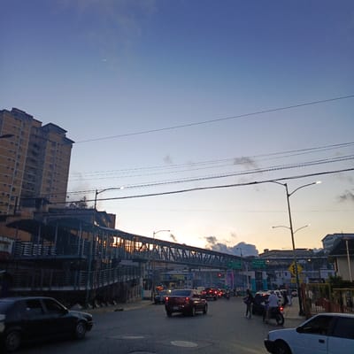 The image depicts a street scene at dusk in Carrizal, Venezuela. A tall, light-orange apartment building dominates the left side of the frame. In the foreground, a dark-colored sedan is captured in motion on the asphalt road. A pedestrian overpass, constructed with metal supports and railings, spans across the road. The sky is a gradient of blue and orange, with clouds visible near the horizon. Several cars are on the road, some of which appear to be in traffic. A motorcycle with two riders is also present. Streetlights are illuminated, and traffic signs are visible, including one indicating directions to "LOS TEQUES," "TEJERIAS," and "LA VICTORIA." The overall environment suggests a busy urban area during twilight.