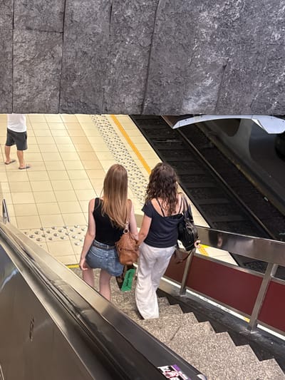 In Molenbeek-Saint-Jean, two women stand on a subway platform. The woman on the left has long, curly brown hair and wears a dark blue tank top with a black backpack. The woman on the right has long blonde hair and is dressed in a black t-shirt and denim shorts, carrying a brown leather purse. Both women are facing away from the camera. A person's legs are visible in the lower left of the frame, wearing white shorts and dark sandals, suggesting another individual is present. The platform is tiled in a light cream color with a decorative yellow stripe at the edge. Visible on the platform is a green shopping bag with the text "lush.com". In the background, a subway train is partially visible, along with stairs and railings, indicating the setting is a metro station. The overall lighting suggests it is daytime, and there are no clear indicators of the weather or the emotional state of the subjects.