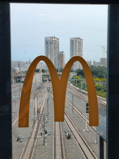 The image is a photograph taken from a window, looking out onto a cityscape and railway tracks. The foreground is dominated by the iconic golden arches of the McDonald's logo, partially obscuring the view. Beyond the arches, a complex network of railway tracks spreads out, with overhead power lines indicating an electrified system. In the middle ground, there are several modern high-rise buildings, typical of urban development. Further in the background, more buildings and some greenery are visible. The sky is a pale blue with a few scattered clouds. The presence of the McDonald's logo suggests this is likely a location visible from a McDonald's restaurant in Harbin, China, potentially overlooking a railway station or yard. There are no discernible people or specific actions captured in the frame. The environment appears to be an urban setting with significant infrastructure.
