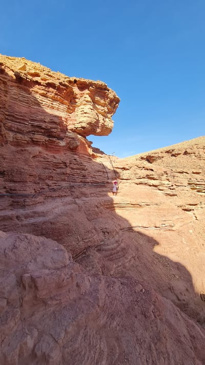The image shows a person on a via ferrata in a rocky desert environment, possibly in the city of Hevel Eilot Regional Council, Israel. The person is wearing a light-colored outfit and appears to be ascending a cliff face equipped with metal rungs and cables. The rock is predominantly red/brown in color and the sky is clear blue. There is a rock formation on the left and a shadow cast on the cliff face to the right.