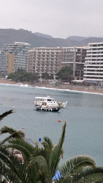 The image displays a coastal view in Podbadac, Montenegro, under an overcast sky. In the immediate foreground, dark green palm fronds partially obscure the lower left and right corners. The midground features a body of calm, teal-colored water. Two boats are anchored: a larger white passenger vessel with the name "TRITON II" visible on its side, and a smaller, dark-hulled boat with white vertical stripes and multiple fenders positioned to the left of the larger vessel. An orange buoy floats prominently in the lower right, with another orange and a blue buoy visible further back. A partially submerged floating platform is also visible. In the background, gentle waves break along a sandy beach. Behind the beach, a densely packed row of multi-story buildings extends horizontally; many exhibit exposed concrete frames, indicating ongoing construction. A building on the far left displays a distinctive gold and yellow patterned facade. Green foliage and structures consistent with beachfront establishments are visible between the beach and the buildings. Distant, forested mountains rise behind the urban structures, fading into the uniformly light gray sky. No individuals are visible within the scene.