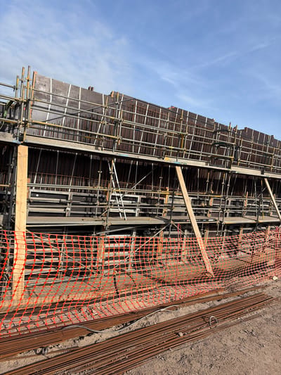 This is a daytime, outdoor photograph of a construction site, likely depicting the preparation for pouring concrete for a vertical structure. The image showcases the skeleton of a reinforced concrete wall or similar element, with various construction materials and temporary structures visible under a clear blue sky.

The central subject is a tall, dark-colored formwork structure, supported by extensive metal scaffolding. Inside the formwork, a dense network of rusty reinforcing steel bars (rebar) is arranged, forming the reinforcement cage for the concrete. The scaffolding consists of numerous metal pipes and clamps, providing structural support and access. Several wooden planks and braces are integrated into the formwork and scaffolding system.

To the right of the main structure, the ground is composed of disturbed brown earth and dirt. Piles of loose rebar rods are laid out on the ground, ready for use. An orange plastic safety netting, acting as a barrier, is prominent in the foreground on the right. The text "WATT" is visible as a logo on this safety netting.

The sky, occupying the upper left portion of the image, is a bright blue with a few wispy white clouds, indicating clear weather. The scene is well-lit by sunlight, which casts shadows, particularly on the textured safety netting and the rebar. No people are visible, but the presence of the materials and structures indicates ongoing construction activity.