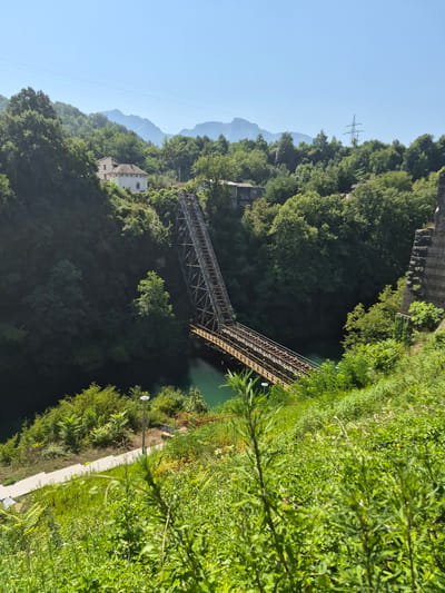 The image shows a view of the historic railway bridge in Jablanica, Bosnia and Herzegovina, spanning across the Neretva River. The bridge, a prominent historical landmark from World War II, is constructed of steel and wood. It is situated within a lush, green, and mountainous landscape. The river below is a vibrant turquoise color. The foreground is dominated by dense green vegetation, with a pathway and stairs leading down towards the river on the left side. A solitary lamppost is visible along the pathway. In the background, several buildings are nestled among the trees on the hillside. The sky is clear and blue, with distant mountains shrouded in a light haze. A power pylon can be seen on a ridge in the far background.