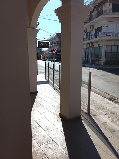 The image depicts a street scene in Λεχαινά, Greece. It is taken from under a covered walkway with white columns and archways, providing a partial view of the street. Sunlight streams in, casting long shadows from the columns onto the tiled walkway.

Across the street is a building with balconies, air conditioning units, and a sign that reads "VASSILIS PANTAZIS AGROFRUT TEL: 26230 22561". Adjacent to the walkway is a glass barrier, possibly for an outdoor seating area. In the distance, a street sign for "ΔΡΟΣΕΛΗ Restaurent Cafe Bar" and a no left turn sign are visible, along with several cars parked along the side of the street.