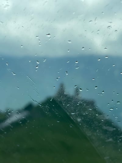 The image depicts a view through a rain-streaked window, with water droplets distorting the scene outside. In the background, a blurred landscape includes rolling green hills and a body of water, likely a lake or a distant river. A prominent, out-of-focus structure, possibly a church or a fortified building with spires, is visible against a cloudy sky. The overall impression is of a moody, atmospheric day in a natural setting. The provided information states that this is the city of Gergeti, Georgia.