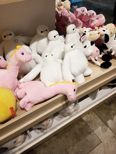 The image displays a retail merchandise arrangement of numerous plush toys on a multi-level shelving unit within a commercial establishment. The upper and middle shelves are densely populated with a variety of stuffed animals. Dominating the arrangement are numerous white, rotund, humanoid-shaped plush figures, resembling Baymax characters, approximately two dozen of which are visible in various upright and reclining positions. Prominently featured in the foreground are two pink, long-necked dinosaur-like plush toys; one stands upright on the left, and another lies horizontally across the front edge of the middle shelf. Other plush subjects include various pink animal figures, possibly pigs or hippos, and multiple cream-colored and black-and-white spotted cow-like figures, primarily visible in the background and on the right side. A partial view of a yellow, spherical plush item is visible on the far left foreground. The shelving unit consists of light-colored wooden planks supported by white uprights, with white open compartments visible underneath the main display surface, some containing smaller plush items encased in clear plastic bags. The floor visible beneath the shelving unit is a dark, mottled concrete or tiled surface. This scene is located within a commercial space in Tân Kỳ Commune, Vietnam.