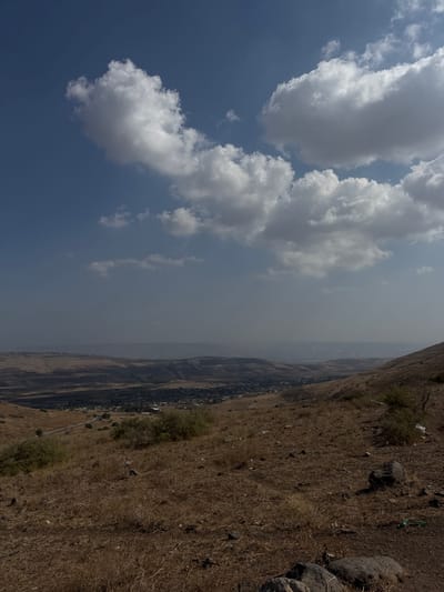 The image displays an expansive, elevated landscape view within the Galil Tachton Regional Council, Israel. The upper portion of the frame features a blue sky dotted with scattered cumulus clouds, presenting varied shades of white and grey. The foreground consists of a sloping, arid hillside covered predominantly with parched golden-brown and light brown grasses, dry earth, and scattered dark grey rocks. Sparse, low-lying green-brown shrubs are visible across this dry terrain. No individuals, animals, or active subjects are present. In the midground, the land descends into a wide valley. A narrow, dark-colored road is visible winding through the mid-left section of the landscape. A compact settlement or community, characterized by clusters of dark-roofed structures, is situated within the valley. The background shows successive layers of distant mountains and hills receding into a pale, bluish atmospheric haze, extending to the horizon.