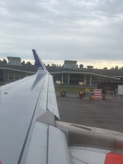 The image shows an airplane wing in the foreground, with a view of an airport terminal in the background. The terminal building has large glass windows and a sign that reads "Are Östersund Airport" and "Welcome to Are. Home of Ostersund." There are some vehicles and equipment on the tarmac, including a small yellow loader and a red and white barrier. The sky is overcast with some patches of sunlight, suggesting it is either early morning or late afternoon. The overall scene depicts an airport setting, possibly during a boarding or deplaning process. The SAS logo is visible on the tail fin of the airplane.