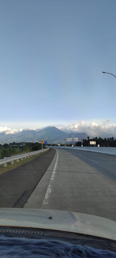 The image depicts a scenic view of a highway in Baksukon, Indonesia. The perspective is from inside a vehicle, looking forward along the paved road. The highway curves gently to the right, with two lanes visible, separated by white markings. To the left of the road, a metal guardrail runs along the shoulder, beyond which is a grassy area and some vegetation. In the distance, a large mountain range dominates the horizon, with clouds partially obscuring its peaks. Several signs are visible along the roadside, including a directional sign with a yellow arrow and a blue sign indicating "Truk Ke Kiri". The sky is clear and a pale blue, with a few scattered clouds near the mountains. A street lamp can be seen on the right side of the image, extending into the sky. The foreground shows the dashboard of the vehicle, covered with a blue fluffy material. The overall impression is of a peaceful journey on a highway with a beautiful natural backdrop.