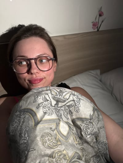 The image displays a young adult female positioned in a bed in Ercolano, Italy. She has light skin, dark hair, and wears round-framed eyeglasses. Her light-colored eyes are directed towards the upper right, and her lips are slightly parted, exhibiting a glossy texture. Her upper body is partially obscured by a grey textile featuring an elaborate baroque pattern with gold and dark grey accents. To her right, white pillows are visible. Behind her, a wooden headboard is present, above which a white wall displays a graphic of two pink flowers with dark stems. The scene is dimly lit, with shadows prominent on the left side of the subject and along the headboard.