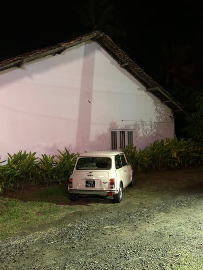 A white Mini Cooper is parked on a gravel and grass area next to a white building. The car is viewed from the rear and right side. A license plate on the back of the car reads "483 7955". The building has a sloped roof with a thatched edge and a small window. Green foliage lines the side of the building and the driveway. The scene appears to be outdoors at night. The lighting is dim, casting shadows from the foliage onto the building. The overall mood is quiet and still. The image was taken in Talpe, Sri Lanka.