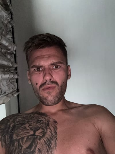 The image shows a man's chest and face, with a large lion tattoo on his left chest. He has short, dark hair and a beard. His expression is a slight grimace. To the left of the man is a draped curtain with a floral pattern. The background is a plain, light-colored wall. The lighting is dim, casting shadows on the man's face and chest. The scene appears to be indoors, likely in a bedroom or living area, and is not specific to any location in Sesto Calende, Italy.