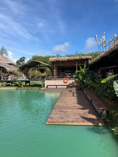The image shows an outdoor scene with a body of turquoise water in the foreground, a wooden boardwalk extending into it, and several thatched-roof structures set back from the water. The structures appear to be part of a resort or restaurant, with some featuring open-air seating and bamboo detailing. Lush green vegetation, including large-leafed plants and yellow flowers, is visible around the buildings. A life preserver is hanging on one of the bamboo walls. The sky is blue with scattered white clouds, indicating fair weather. The location is identified as Tambon Pa Kae Bo Hin, Thailand. There are no people visible in the image.