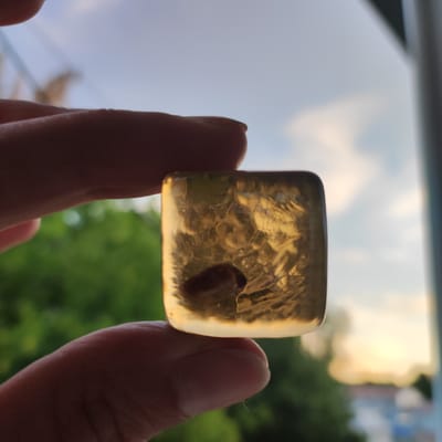 A close-up shot shows a person's fingers holding a small, translucent, square-shaped object. The object appears to be amber or a similar resin, with an inclusion visible inside, possibly an insect or plant matter. The object is lit from behind, allowing light to pass through and illuminate its textured interior. The background is blurred, suggesting an outdoor setting.
The blurred background reveals the soft green foliage of trees and a sky with hints of blue and warm, golden light, indicative of either sunrise or sunset. A vertical dark shape on the right side of the frame could be a window frame or railing. The overall lighting and colors suggest a serene, natural setting. The focus is sharp on the object and the fingers holding it, while the background is softly out of focus, creating a shallow depth of field. There is no visible text in the image. The scene appears peaceful, with the warm light creating a gentle ambiance. The location context provided is Kineshma, Russia.