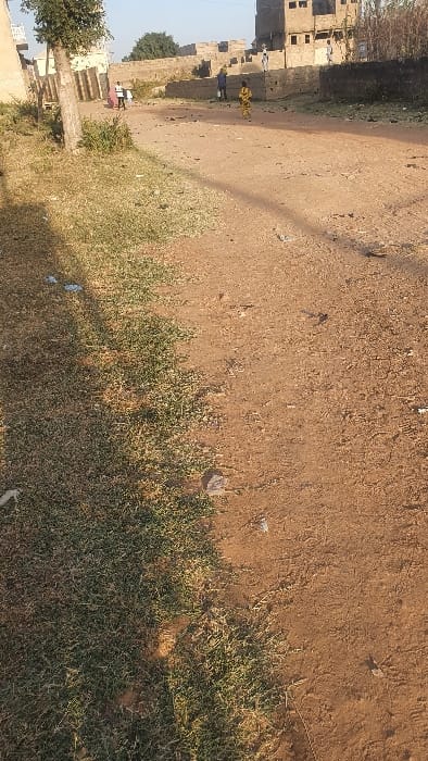 The image displays an unpaved, reddish-brown dirt thoroughfare within the city of Danbare, Nigeria, under bright daylight. The left foreground features a strip of dry, green-brown grass, with bare earth and scattered small stones and debris covering the remainder of the foreground and midground. A dark shadow extends diagonally across the lower-left portion of the frame.

In the midground, five individuals are present. On the left, one person, clad in a light top and dark trousers, walks away from the viewer, carrying a large, light-colored rectangular object, consistent with a mattress or panel, over their left shoulder. To the right of center, another individual, dressed in a patterned yellow and black garment, moves towards the right side of the image. Further into the background, a person in blue clothing stands near a wall, and two additional figures in light-colored attire are visible among the background structures.

The background consists of several buildings, primarily constructed from concrete blocks. A multi-story structure on the far right appears partially completed or in disrepair. Stone or concrete walls define property boundaries. Sparse trees and bushes are present on the periphery of the scene. No explicit interactions between individuals are discernible.