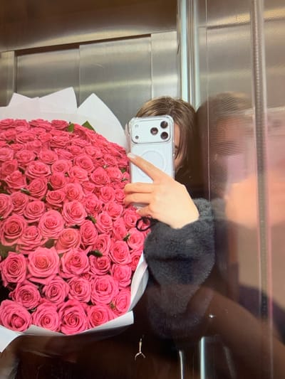 A person is taking a selfie in a reflective surface, likely an elevator or a polished wall, holding a large bouquet of pink roses. The bouquet is the dominant object, filling the lower half of the frame. The person's arm and hand are visible, holding a smartphone to capture the reflection. The person is wearing a dark, fuzzy-textured jacket. The background appears to be a sterile, metallic environment with horizontal lines suggesting a structured surface. The lighting is bright, reflecting off the surfaces. The overall impression is one of a moment captured, possibly a celebration or a gift of flowers. There is no visible text in the image.