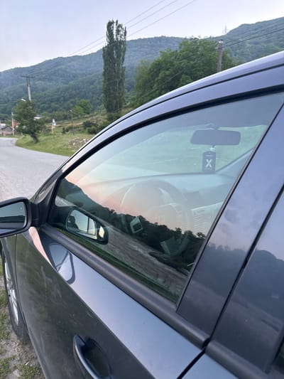 This is a photograph of the interior and exterior of a dark-colored car, taken from a side angle. The image is oriented vertically, with the sky appearing at the bottom and the ground at the top.
Inside the car, visible through the windshield, is the steering wheel and dashboard. A car air freshener, labeled "X ACTIVE," hangs from the rearview mirror. The windshield also reflects the sky, showing a gradient of colors suggesting either sunrise or sunset.
The exterior of the car shows the side door and window. Scratches and dust are visible on the car's surface. A side mirror is attached to the door.
The scene outside the car, reflected in the window and visible beyond the car, depicts a rural landscape. There are green, rolling hills covered in trees, and a power line pole with wires runs through the scene. The sky is overcast, and the light suggests it might be late afternoon or early evening. The location is identified as Каменномостский, Russia. There is no visible activity or event taking place; the image appears to be a still shot of the car and its surroundings.