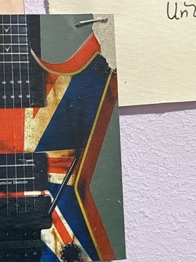 A close-up shot captures a section of an electric guitar with a Union Jack design. The guitar features black pickups and fret markers with white inlay. The Union Jack pattern is weathered and distressed, with visible paint chips and a torn edge on a sticker or decal. To the right of the guitar, a cream-colored surface is visible, with handwritten text that appears to say "Un". Below this, a textured, pale purple wall is seen. The image is tightly cropped, focusing on the guitar's body and a portion of the wall. The specific location in the city of Jrarat, Armenia, cannot be determined from the provided image.