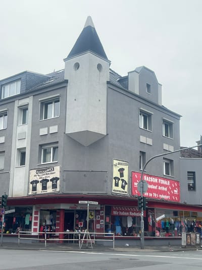 This image captures a street view in Herne, Germany, focusing on a multi-story building with a distinctive turret. The building's facade is primarily gray, with white accents around the windows and a light gray projecting section with a dark conical roof at the top. Below the turret, two circular windows are visible, resembling eyes, giving the structure a somewhat anthropomorphic appearance.
The ground floor of the building houses a "PROFI SHOP," which appears to be a retail store, possibly selling sports apparel, indicated by "PUMA" branding and images of t-shirts on signage. Red awnings extend over the storefront windows, which display various items of clothing and merchandise. A prominent banner with "SAISON FINALE" (Season Finale) and "% Auslauf Artikel" (Outlet Items) suggests a sale is in progress, with discounts up to 70%. Another sign reads "Wir bringen Ihre Ideen auf's Shirt" (We bring your ideas onto a shirt), referencing a "FLOCKSERVICE" (Flocking Service).
A traffic light stands to the left of the building, and a metal railing separates the sidewalk from the street. The sky is overcast, suggesting a cloudy day. The street itself appears to be a typical urban road with pavement and road markings. There are no clear individuals visible interacting with the scene, though some figures may be dimly observed within the shop windows. The overall environment is that of a commercial street in a German city.