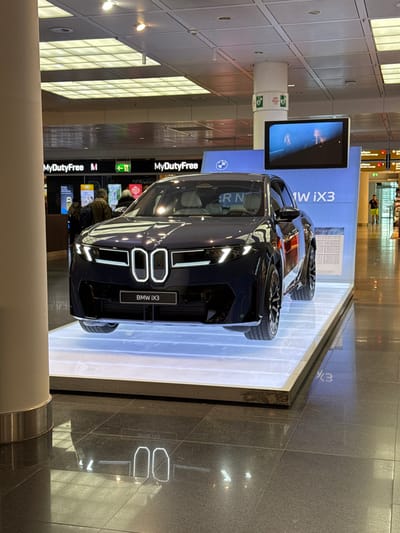 A dark-colored BMW iX3 electric SUV is displayed on a rectangular, illuminated white platform at Munich Airport, Freising, Germany. The vehicle is positioned facing slightly towards the left of the frame, presenting its front fascia, which features an illuminated kidney grille outline and activated headlights. A black panel below the grille prominently states "BMW iX3". Behind the vehicle, a large blue display wall exhibits the BMW logo and "BMW iX3" branding. An overhead monitor, suspended from a white structural pillar, displays dynamic visual content. The dark, polished tiled floor reflects the illuminated elements of the display, including the car's grille lights and the platform's ambient glow. In the background, multiple signs are visible, including "MyDutyFree M" to the left and "Lufthansa Lounge" further to the right, adjacent to an exit sign. Several unidentifiable individuals are observed moving through the terminal; some are near the retail establishments on the left, and one male subject in a yellow shirt is standing further to the right. The environment is an brightly lit indoor airport terminal characterized by a grid of overhead artificial lighting. A large, light-colored structural pillar is partially visible in the left foreground of the image.
