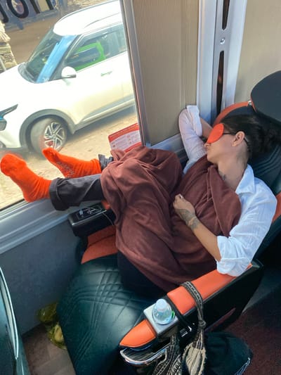 A woman is asleep on a bus, wearing an orange eye mask and covered with a rust-colored blanket. Her feet, clad in orange socks with white grips, are propped up on the window ledge. A white shirt is visible beneath the blanket. The bus interior features black and orange seating. Outside the window, a white car is parked. The scene is set in Liên Nghĩa, Vietnam.