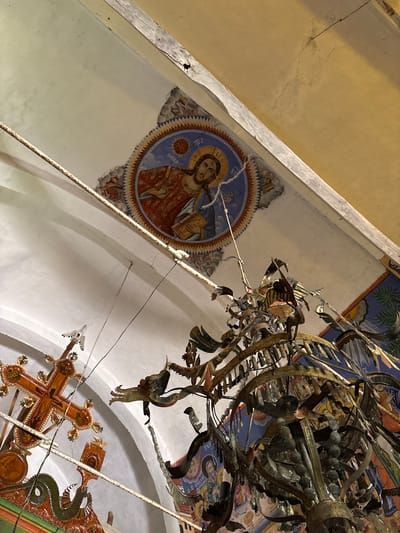 The image shows the interior of the church of St. George in Gorno Uino, Bulgaria. A religious fresco of Jesus is painted on the upper left wall inside a circular frame. To the right, a large, ornate metal chandelier dominates the view, with hanging decorations in the shape of birds. A wooden cross with decorative elements is also visible in the lower right corner. The walls are painted white, with an arched architectural design. The overall lighting suggests an interior space.