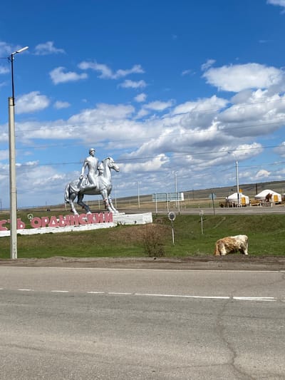 The image shows the entrance sign to the city of Ust'-Ordynskii, Russia. A silver sculpture of a horse and rider stands next to the sign with the city name in Russian. The sky is blue with white clouds. A grassy area is adjacent to a road, and a dog is lying in the grass. Utility poles and lines are visible, as well as some buildings and structures in the background.