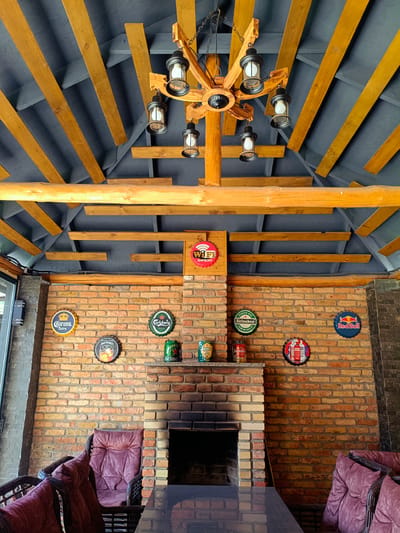 The image depicts an outdoor covered seating area, possibly a patio or gazebo, with a rustic and industrial aesthetic. The ceiling is constructed with dark grey panels and exposed wooden beams, supporting a rustic chandelier with multiple lantern-style lights. The walls are made of exposed orange and yellow brick.

Decorations on the brick wall include several circular metal signs, resembling bottle caps, with various brand logos such as Corona Extra, Jack Daniel's, Heineken, and Red Bull. There is also a wooden sign with a "Wi-Fi OPEN" graphic. A fireplace with a dark hearth is built into the brick wall.

In the foreground, there are plush, upholstered chairs with dark wicker frames, positioned around a dark, reflective table. The chairs are upholstered in a deep purple fabric. The overall atmosphere suggests a relaxed, casual establishment, likely a bar or restaurant with an outdoor seating area. The image is taken from a low angle, looking upwards towards the ceiling and fireplace. There are no people visible in the image. This establishment is identified as being located in the city of Kok Jar, Kyrgyzstan.