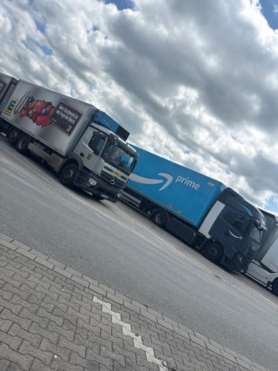 The image captures a line of parked delivery trucks in Beckum, Germany. The trucks are parked on a paved roadside with a brick-paved sidewalk. The sky is overcast with heavy clouds. An Amazon Prime truck is visible with its signature blue trailer and logo. An EDEKA truck is also present. Text on the EDEKA truck reads: "Guter Geschmack ist eine Frage der Beere." The license plate on the EDEKA truck reads "WES ED 310". Other truck details include "FRIGOBLOCK" on top of the truck, and "IVECO" on the side of the Amazon Prime truck.