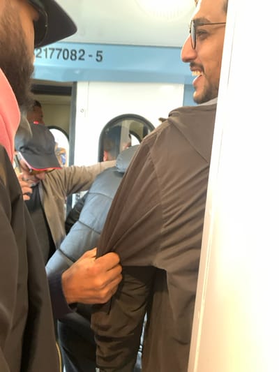 Several people are inside what appears to be a laundromat. A man with glasses is seen smiling. Another person is partially visible wearing a black hat. Washing machines with round windows are visible in the background. A person is holding the fabric of a gray jacket or coat. Number 2177082-5 is written on a light blue surface.