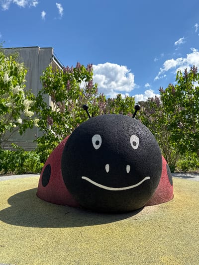 The image shows a large, stylized ladybug sculpture on a yellow playground surface. The ladybug is black with red markings and a smiling face. It is located in an outdoor setting with trees and a building visible in the background. The sky is blue with scattered clouds. Based on the location information, this is likely in Jaunmārupe, Latvia.