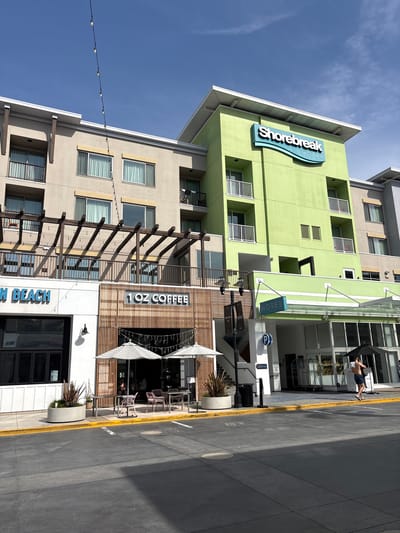 A daytime view captures a lively street scene in Huntington Beach, United States of America, featuring a multi-story hotel and a coffee shop under a clear blue sky with sparse clouds.
The prominent structure is a vibrant lime-green building housing the "Shorebreak HOTEL," with its name displayed in large blue letters. A smaller blue "HOTEL" sign hangs above the main entrance, which is sheltered by a glass canopy. Below the green hotel, another building features a facade with vertical wooden slats, clearly marked with "1 OZ COFFEE" and a partial sign reading "N BEACH."
On the street level, a person is visible near the hotel entrance, holding a large black umbrella, possibly for shade given the sunny weather. A luggage cart is also present. In front of the coffee shop, outdoor seating with tables, chairs, and two large white-and-grey striped umbrellas suggests an inviting al fresco dining area. The street itself is asphalt with a distinct yellow curb line. Other details include balconies on the buildings, decorative string lights stretched across the scene, and a blue circular "P" sign, likely indicating parking.
The bright lighting suggests it is either morning or early afternoon. The overall impression is that of a modern, bustling urban environment, typical of a coastal resort town. No specific emotions are discernible from the image.