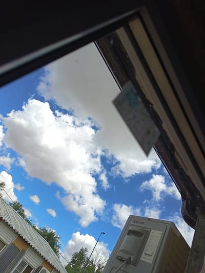 The image captures a skyward view from what appears to be an outdoor setting, possibly in the village of Seitovka, Russia, given the context. The sky is a vibrant blue, populated with fluffy white cumulus clouds. In the foreground, a portion of a corrugated metal roof of a building is visible, with white siding beneath. Trees with green foliage are scattered in the background. To the right, part of a dark, possibly metallic structure, like a gutter or overhang, is present. At the bottom right, a gray, rectangular object, likely an outdoor unit of an air conditioner, is positioned. A street lamp with a dark pole is also visible extending into the sky amidst the trees and clouds. The overall scene depicts an outdoor environment with typical village elements.